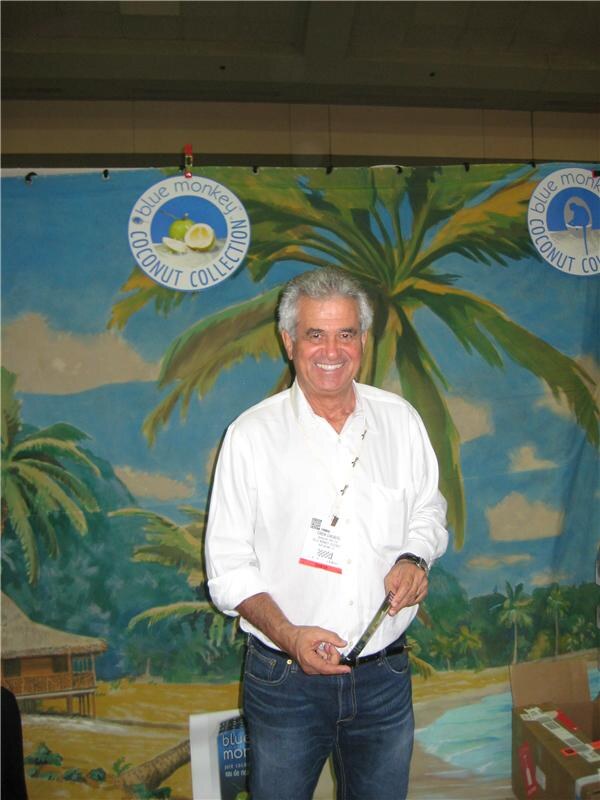A smiling gray-haired man in a white shirt and lanyard stands in front of a tropical palm tree backdrop bearing Blue Monkey Coconut Collection logos.