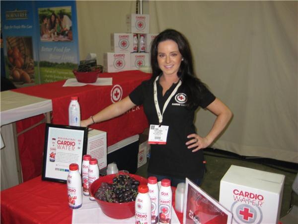 A smiling woman with long dark hair, in a black Cardio Water shirt and lanyard, stands at a red-draped table with Dox bottles, a bowl of grapes and a Cardio Water box. A Born Free banner is behind her.