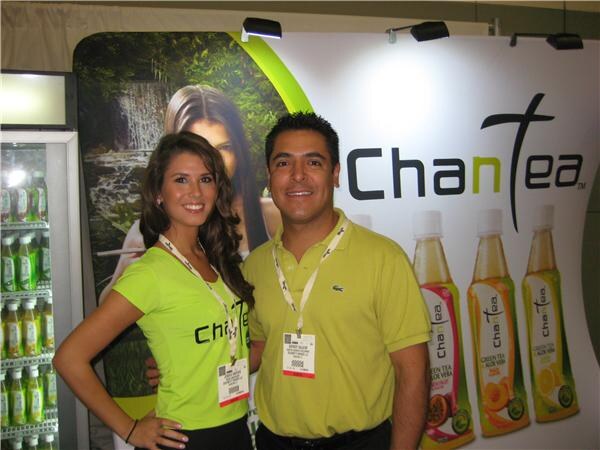 A smiling woman in a green ChanTea shirt and a man in a green Lacoste polo stand together in front of a ChanTea banner showing bottled green tea drinks.
