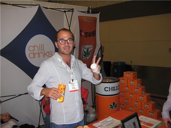 A man with glasses holds an orange can and a small cup in front of a Chill Drinks banner, a C Swiss banner, and stacked orange Chillo boxes.