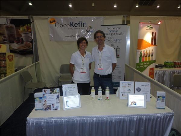 A woman and a man in white shirts stand behind a table with CocoKefir bottles and info cards, under a CocoKefir banner, with another beverage banner at right.