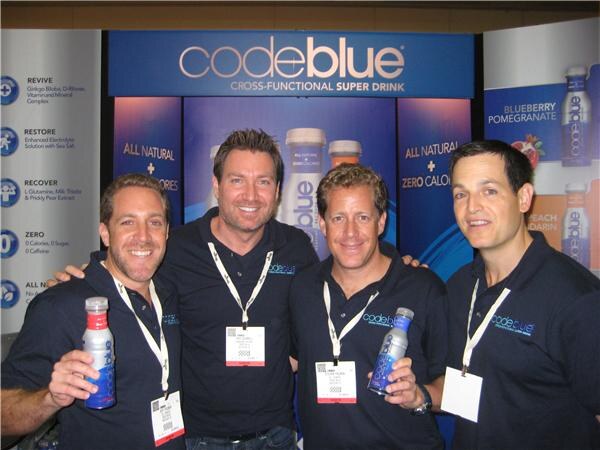 Four smiling men in navy Code Blue shirts and lanyards stand together in front of a codeblue booth backdrop. Two hold bottles of the drink. Banners read All Natural and Zero Calories.