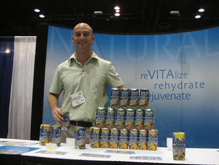 A bald man in a light green shirt and name badge stands behind a table with a stacked pyramid of Vita Coco cartons. A blue backdrop reads 'reVITAlize, rehydrate, rejuvenate'.