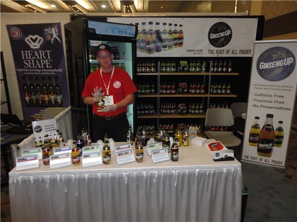 A man in a red shirt and cap, wearing gloves, holds a bottle behind a white-draped table of Ginseng Up sodas. A Heart Shape banner is at left and a Ginseng Up banner at right.