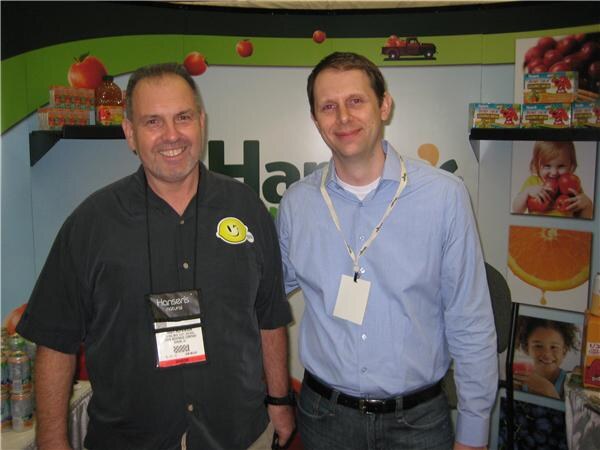 Two smiling men stand side by side in front of a Hansen's Natural booth with juice boxes and fruit photos. One wears a dark shirt with a badge, the other a blue shirt and lanyard.