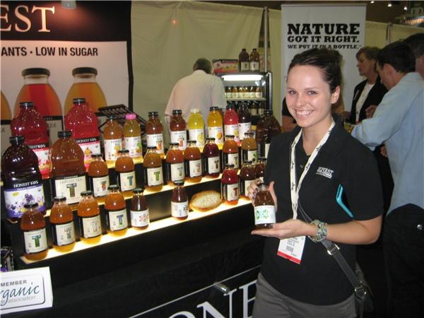 A smiling woman in a black Honest Tea polo holds a bottle in front of a lit shelf display of Honest Tea bottles. A sign reads Nature, Got It Right. Other attendees stand behind her.