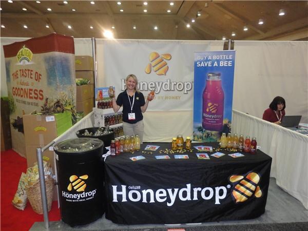 A blonde woman holds up two bottles behind a black table marked Honeydrop. A banner reads Buy a Bottle Save a Bee. Healthy Harvest packaging is at left, and a seated woman works at a laptop.