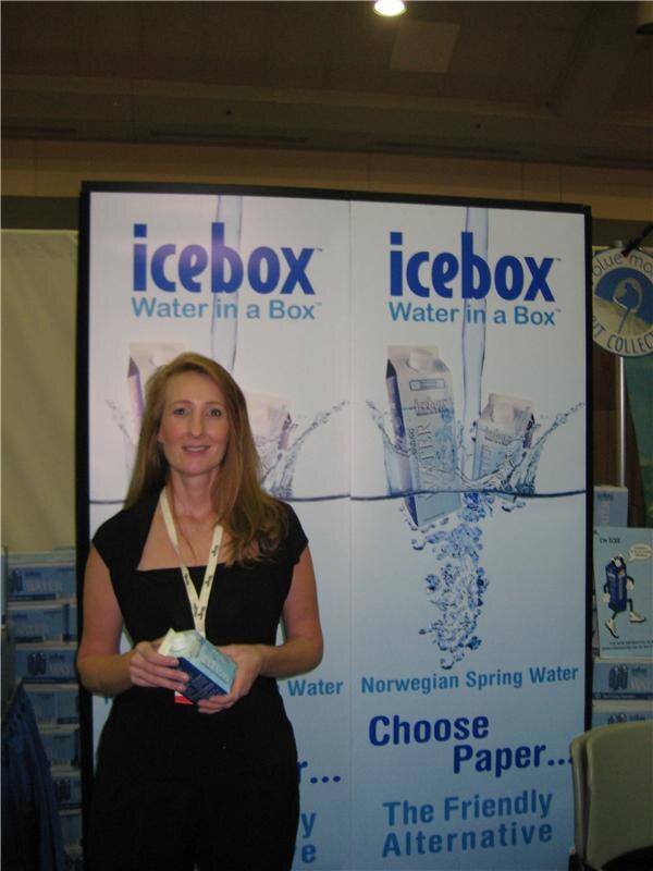 A woman with long hair in a black top holds a small carton, standing in front of an icebox Water in a Box banner that reads Norwegian Spring Water and Choose Paper.