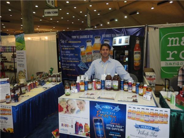 A man in a light blue shirt stands behind a table of Inko's White Tea bottles and cans, with a White Tea Energy banner. A Maine Root booth with green signage is at right.