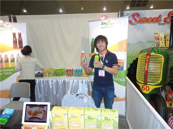 A smiling man in a navy polo and lanyard holds two bottled drinks in a Jayone Foods booth. A woman stands at left near a table of bottles. A Sweet Elena banner and green tractor are behind.