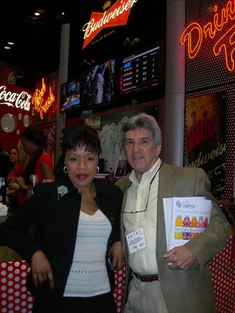 A woman in a black jacket and a man in a tan blazer holding Vitaminwater flyers stand in front of Budweiser and Coca-Cola signs and neon lights.