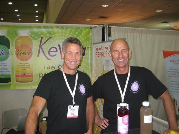 Two smiling men in black T-shirts and lanyards stand side by side in front of a KeVita banner. Bottles of drinks sit on the table in front of them.