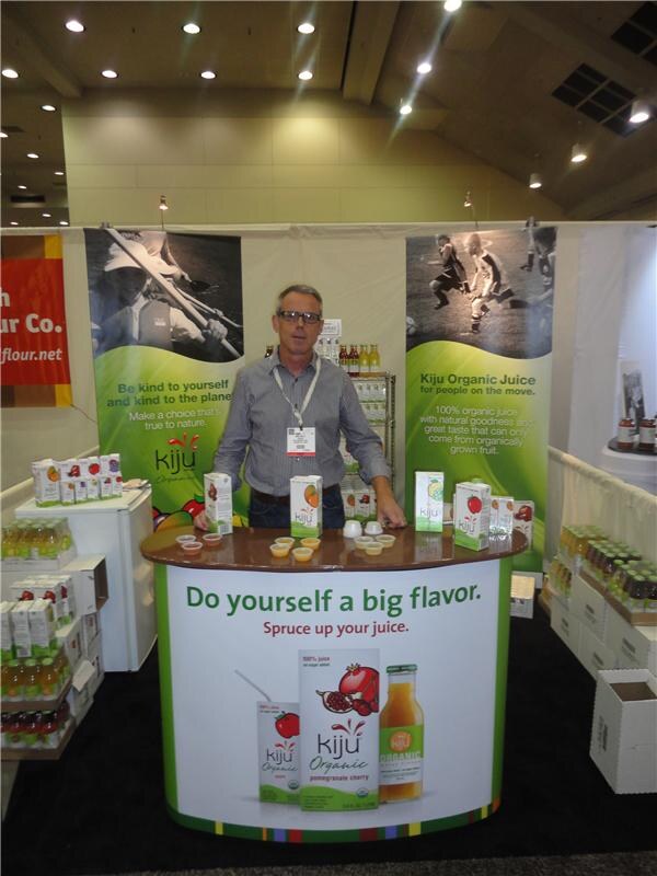 A man in glasses and a gray striped shirt stands behind a Kiju Organic juice counter that reads "Do yourself a big flavor." Juice cartons and sample cups sit on top, with banners behind him.