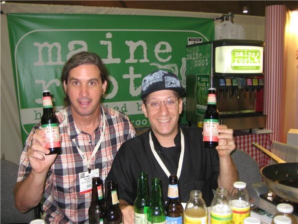 Two men hold up bottles of soda in front of a green Maine Root banner. One wears a plaid shirt and the other a black cap and black shirt. More bottles sit on the table and a fountain is behind them.