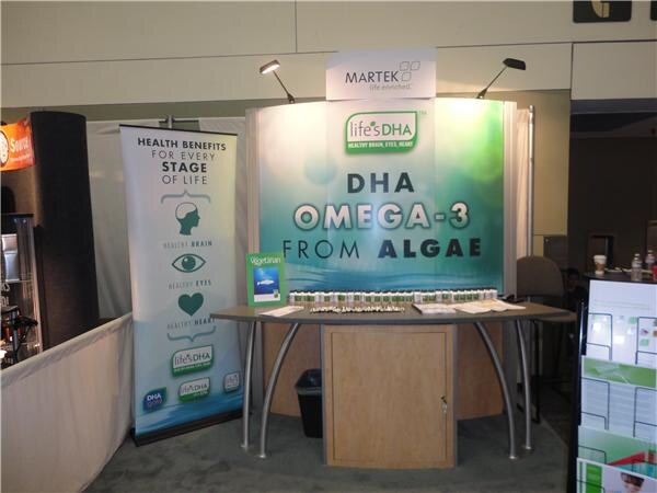 An empty Martek booth with a sign reading "life'sDHA DHA Omega-3 from Algae." A banner at left lists health benefits, and supplement bottles line the curved table.