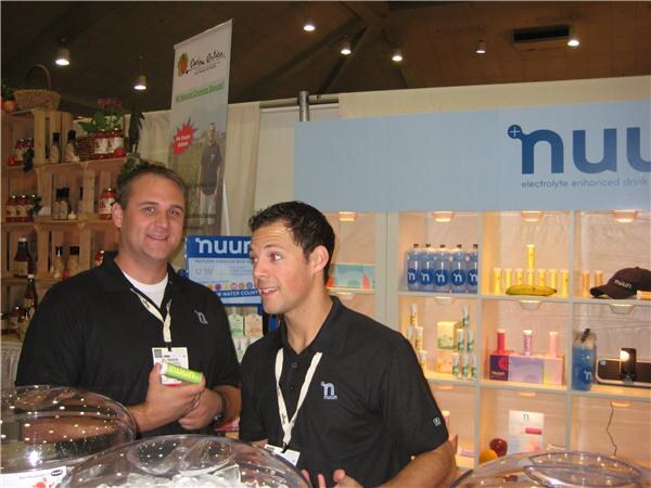 Two men in black nuun polo shirts and lanyards stand at a booth. One holds a green tube. Shelves behind them display nuun electrolyte drink products under a nuun sign.