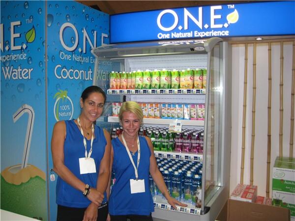 Two women in blue sleeveless tops and badges stand in front of an open refrigerated case of O.N.E. coconut water. An illuminated O.N.E. sign is above, with bamboo at right.