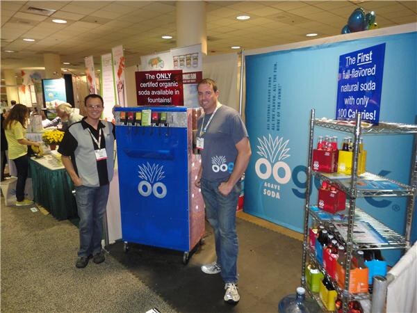 Two men stand beside a blue soda fountain at the Oogave booth. A shelf of bottled sodas is at right, and signs say it is the first full-flavored natural soda with only 100 calories.