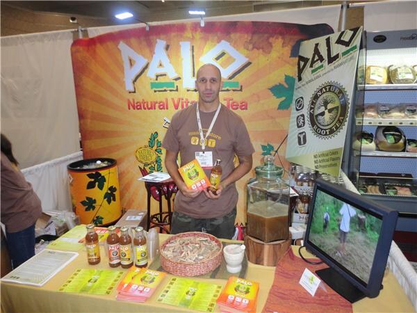 A man in a brown T-shirt and lanyard holds a bottle and a flyer at the Palo booth. Bottles, a glass dispenser, brochures and a monitor sit on the table in front of a Palo banner.