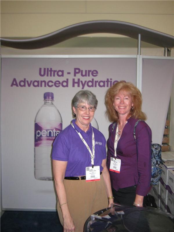 Two smiling women wearing badges stand in front of a Penta backdrop reading "Ultra-Pure Advanced Hydration" with a Penta water bottle. One wears a purple polo, the other a purple blouse.