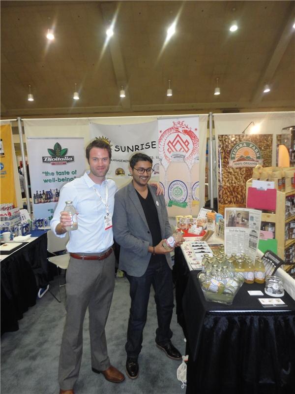 Two men hold glass bottles in a booth with banners for Prometheus Springs, Biotalia and Tierra Farm. A table at right holds bottles of juice and printed materials.