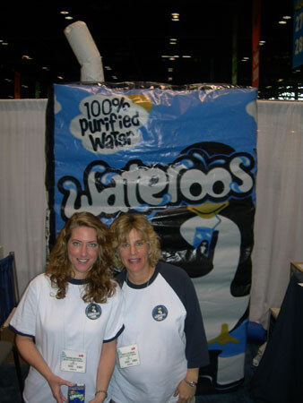 Two smiling women in white and navy shirts with name badges stand in front of a giant inflatable carton reading '100% Purified Water' with a Wateroos penguin logo.