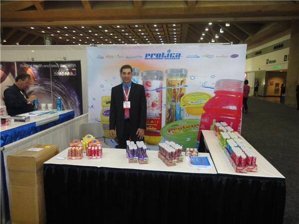 A man in a dark suit stands behind a table of Protica products in front of a Protica Nutritional Research backdrop. Another man sits at a neighboring booth at left.