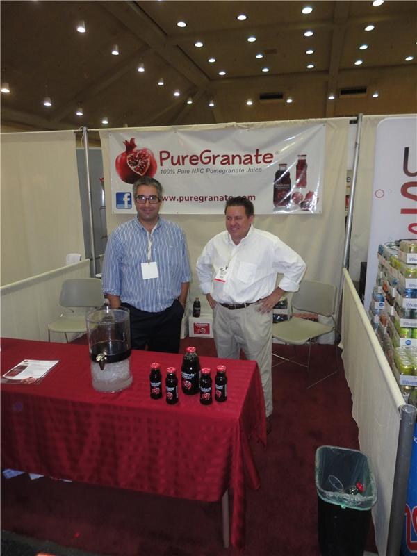 Two men stand behind a red-covered table with bottles of PureGranate pomegranate juice and a drink dispenser, under a PureGranate banner.