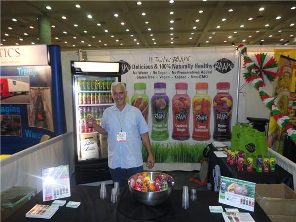 A smiling man holds up a bottle in a RAAW booth. Behind him are a refrigerated cooler and a backdrop of RAAW juice bottles, and a bowl of bottles sits on the table.
