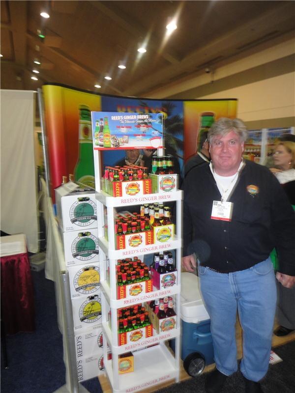 A man in a dark shirt and jeans stands beside a white display rack of Reed's Ginger Brew bottles, with cases of Reed's stacked at left and a colorful backdrop behind.