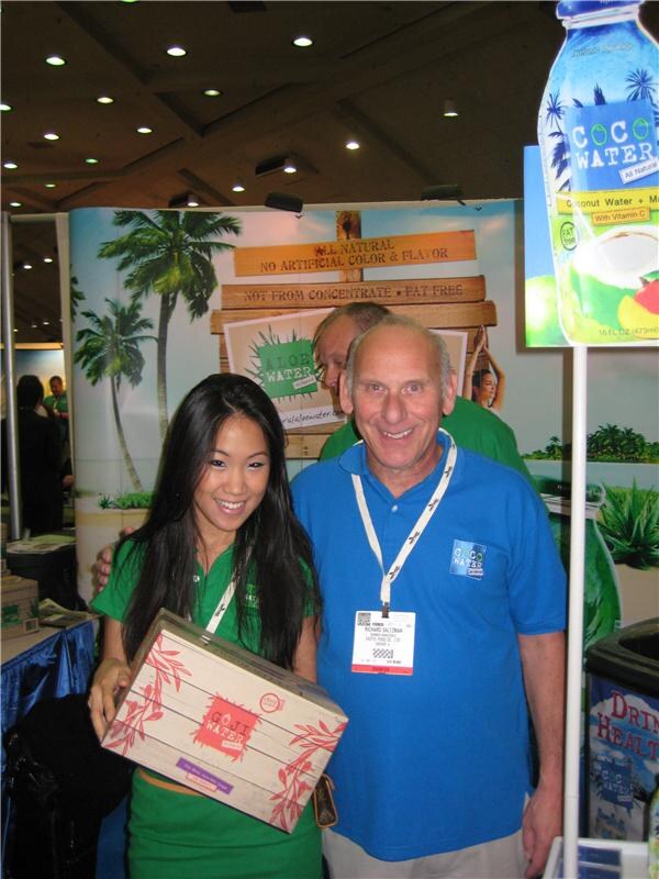 Richard Saltzman, in a blue Coco Water polo and badge, stands smiling beside a woman in green holding a Goji Water box. Coco Water and Aloe Water signage and palm tree graphics are behind them.