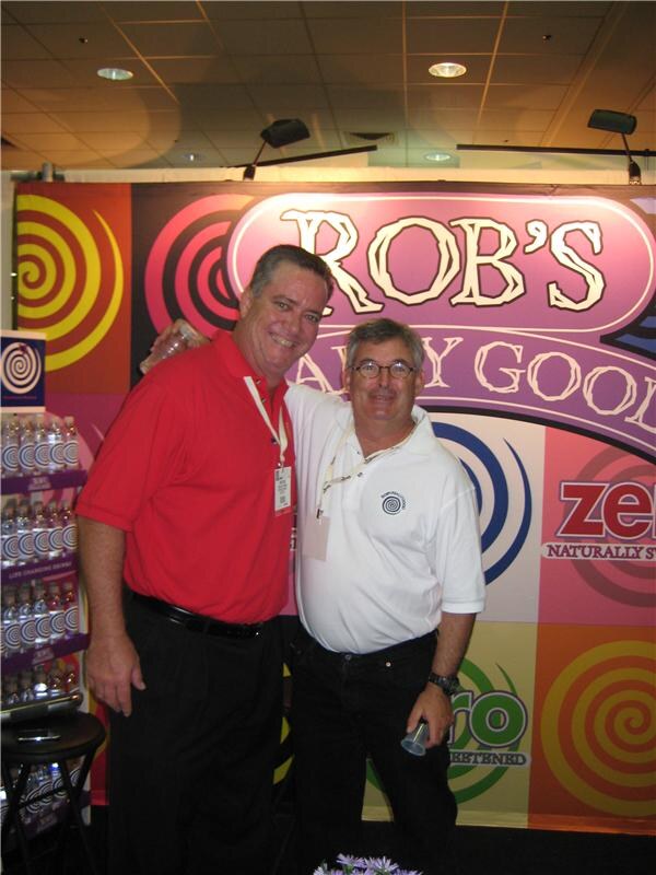 Two smiling men stand close together in front of a Rob's Really Good backdrop with colorful spiral designs and Zero branding. A shelf of bottled drinks is at left.