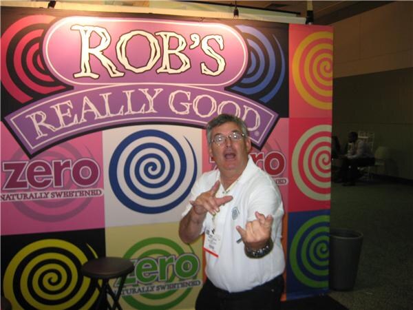 A man in a white polo and glasses gestures playfully with both hands in front of a Rob's Really Good backdrop with spiral designs and Zero signs.