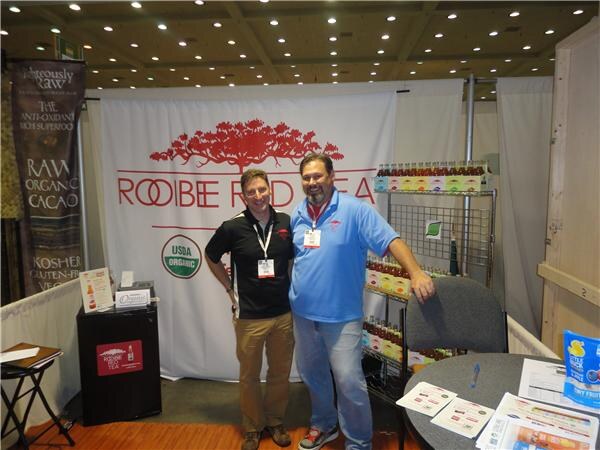 Two men stand side by side in front of a Rooibee Red Tea backdrop with a USDA Organic seal. Shelves of bottled tea are behind them and printed handouts lie on a table.