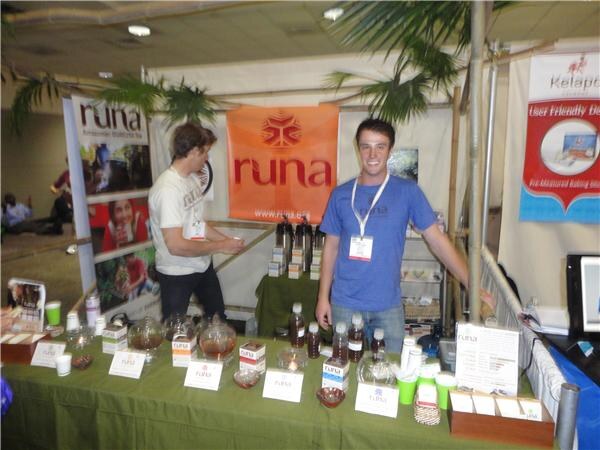 Two men stand behind a green-covered table of Runa tea products and bottles, in front of an orange Runa banner and palm fronds.