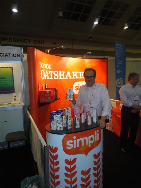 A man in a striped shirt and glasses holds a carton beside a Simpli display with oat drink cartons on a counter, under an orange backdrop reading "Do you Oatshake?"