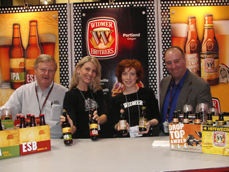 Two men and two women stand behind a counter holding Widmer Brothers beer bottles, with cases of ESB and Hefeweizen in front and Widmer Brothers banners and bottle graphics behind them.