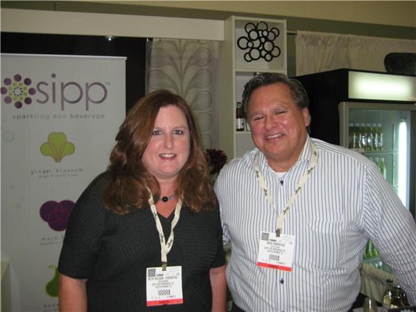 A woman with long brown hair and a man in a striped shirt, both wearing badges, stand together in front of a Sipp sparkling eco beverage banner and a drinks cooler.