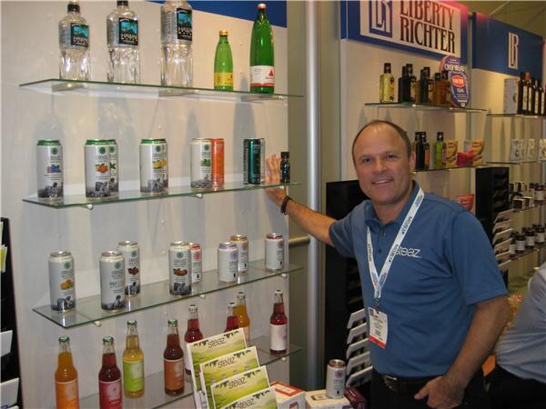A man in a blue Steaz polo shirt rests a hand on a glass shelf of canned and bottled Steaz drinks, next to a Liberty Richter display of bottled products.