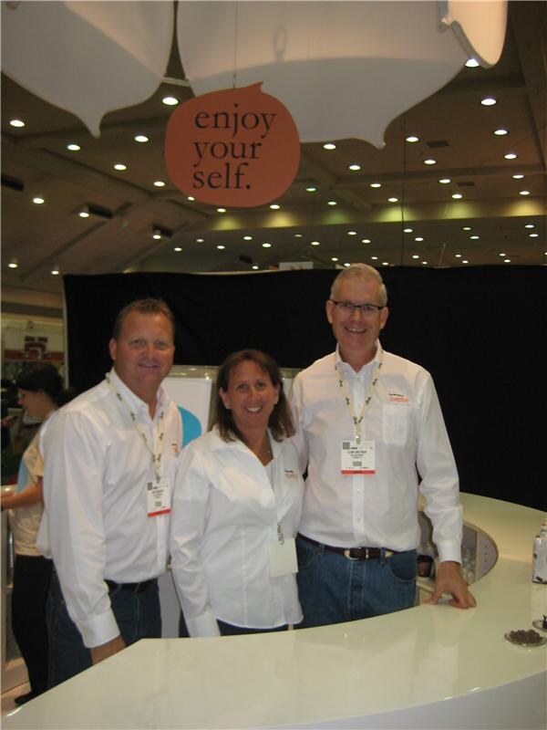 Two men and a woman in white shirts and lanyards stand at a curved white counter under an orange hanging sign reading 'enjoy your self.' at the Svelte booth.