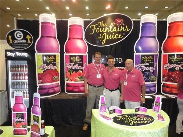 Three men in pink polo shirts stand in front of The Fountains of Juice booth with large bottle cutouts labeled Beetox, Melon and Cherry. A Gigunda Group sign hangs at left.