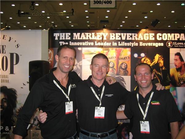 Three smiling men in black shirts and lanyards stand with arms around each other in front of a sign reading "The Marley Beverage Company" with images of drinks.