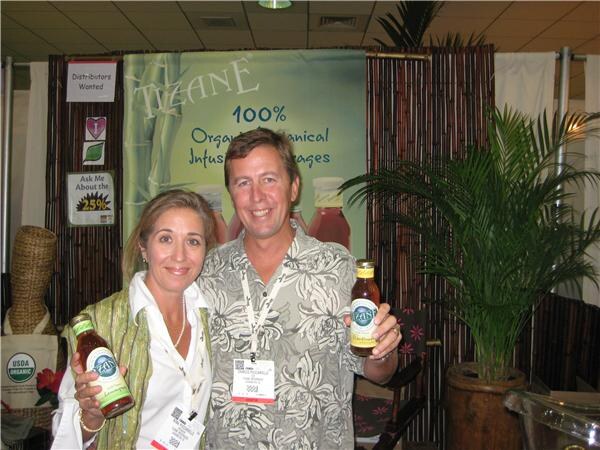 A woman and a man each hold a bottle of Tizane tea in front of a bamboo-style booth with a Tizane banner, a potted palm and a USDA Organic sign.