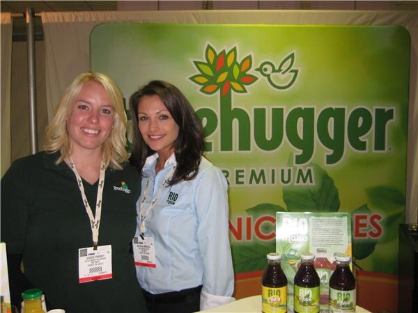 Two smiling women wearing badges stand in front of a green Treehugger Premium backdrop, with bottles of Rio juice on the table in front of them.
