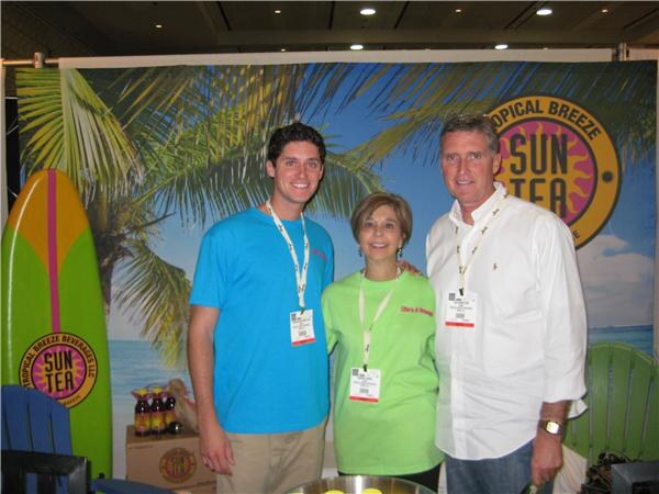 A young man in a blue shirt, a woman in a green shirt and a man in a white shirt stand together in front of a beach-themed Tropical Breeze Sun Tea backdrop with a surfboard.