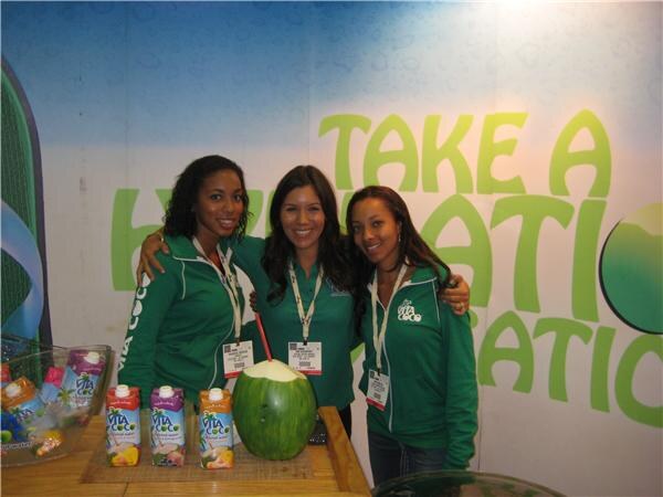 Three women in green jackets stand with arms around each other behind a table holding Vita Coco cartons and a green coconut with a straw, in front of a "Take a Hydration" wall graphic.
