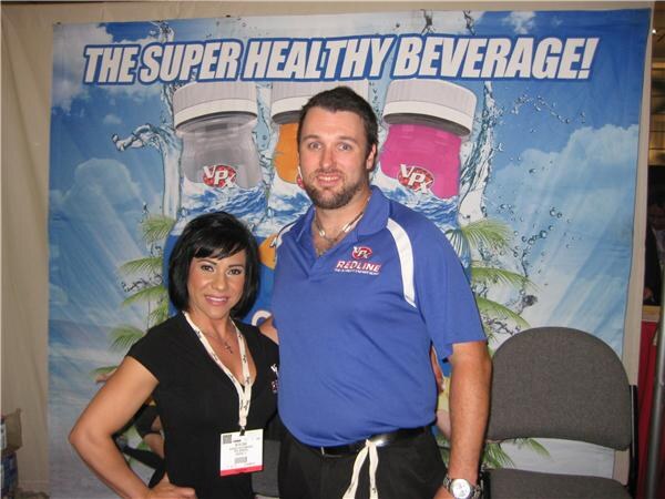 A woman in a black top and a bearded man in a blue Redline polo stand together in front of a VPX banner reading "The super healthy beverage!" showing bottles.