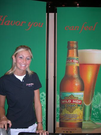 A smiling woman in a dark Stone Mill polo stands beside a large Wild Hop Lager bottle and glass graphic on a green wall reading "flavor you can feel".