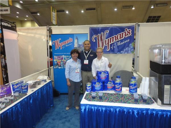Two women and a man stand behind a table with a blue skirt holding bags and bottles of Wyman's of Maine fruit products, with Wyman's banners behind them.