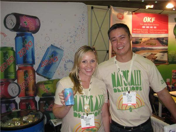 A smiling woman holding a Zevia can and a smiling man stand together in matching green-print shirts in front of Zevia can graphics. An OKF banner hangs behind them.